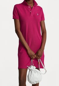 Vestido polo en un vibrante color rosa con cuello y dos botones, que presenta un pequeño logo bordado en el pecho. Acompañado de un bolso compacto blanco.