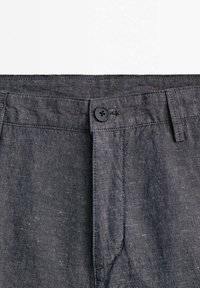 Taille de pantalon gris foncé avec un bouton noir fixé à travers la boutonnière, montrant des passants de ceinture et un tissu texturé.