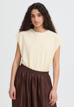 Jonge vrouw met lang krullend haar die een crèmekleurige mouwloze top met hartmotief en een bruine geplooide rok draagt, staand tegen een witte achtergrond.