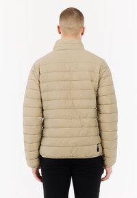 Beige pufferjack met een gewatteerd ontwerp en hoge kraag. Het heeft een gladde textuur en elastische manchetten, gedragen over zwarte broek.