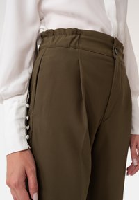 Olijfgroene, op maat gemaakte broek van een textuurstof, met een elastische tailleband en zijzakken. Witte knoopaccenten op de mouw van de blouse.