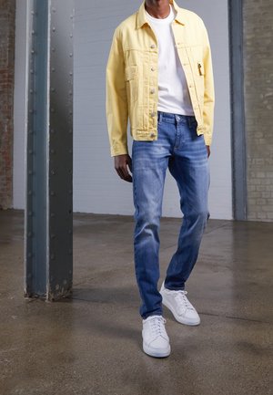 Homme portant une veste jaune clair, un t-shirt blanc, un jean bleu et des baskets blanches, debout à l'intérieur sur un sol en béton près d'une colonne métallique.