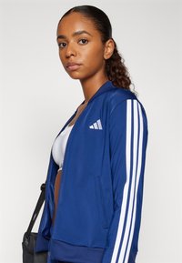 Blå zip-up jacka med vita ränder på ärmarna och dragkedjefickor. Tillverkad av smidigt tyg, med ribbad nederkant och logodetalj.