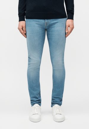 Homme portant un jean skinny bleu clair, une chemise noire à manches longues et des baskets blanches, debout devant un fond uni.