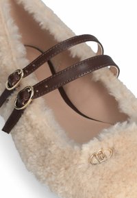 Beige fluffy platte schoen met twee donkerbruin leren banden met gouden gespen en een klein gouden decoratief embleem.
