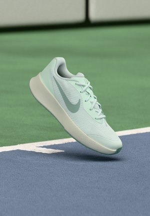 Lys grønn Nike løpesko med lisser, i luften over blå og grønn sportsgulvflate.