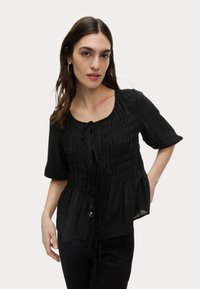 Schwarze Bluse mit Raffung, kurzen Ärmeln und einer Schleifenbindung am Hals, aus leichtem und leicht durchsichtigem Stoff.