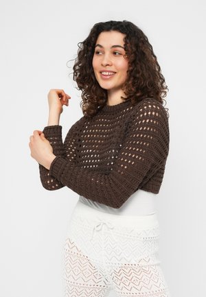 Femme aux cheveux bouclés portant un haut marron en crochet à manches longues et un pantalon blanc à crochet à motifs, souriante et posant de profil.