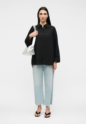 Nő, laza fekete gombos ingben, világoskék, széles szárú, bolyhos farmernadrágban, fekete szandálban, és egy ezüst válltáskát hordva.