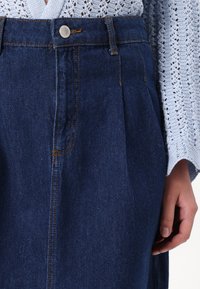 Denim suknja u tamnoplavoj boji s visokim strukom, suptilnim naborima i kontrastnim narančastim šavovima. Ima metalnu dugmad za zatvaranje i prednje džepove.