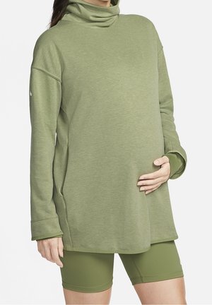 Schwangere Person trägt lockeren olivgrünen Rollkragenpullover und passende enganliegende Shorts, Hand ruht auf dem Bauch.