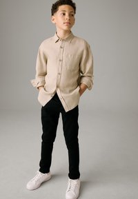 Beiges Leinens-Hemd mit Knöpfen und Brusttasche, kombiniert mit schwarzen Slim-Fit-Hosen und weißen Turnschuhen vor einem neutralen grauen Hintergrund.