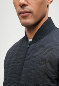 Blouson bomber matelassé noir avec col côtelé et fermeture éclair. Présente un petit logo Lacoste noir sur la poitrine. Tissu texturé.