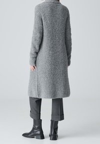 Persona vestida con un cardigan largo texturizado gris, pantalones oscuros grises con puños y botas de tacón negro hasta el tobillo, de pie contra un fondo sencillo.