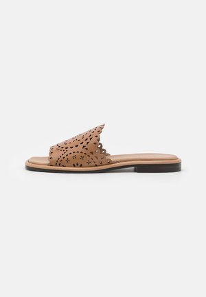 Sandale mule en cuir fauve avec bords festonnés et motif floral ajouré sur la tige, semelle plate noire, vue de côté sur fond blanc.