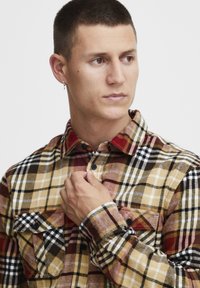 Camisa de franela a cuadros en una mezcla de tonos amarillos, marrones, rojos y negros. Presenta dos bolsillos frontales, cierres de botones y un diseño de cuello clásico.