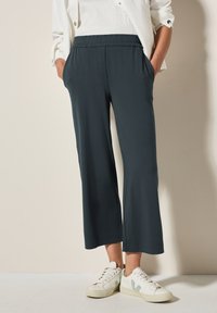 Pantalons cropped vert foncé avec une taille élastique et des poches, fabriqués en tissu lisse. Assortis à des baskets blanches avec des accents verts.
