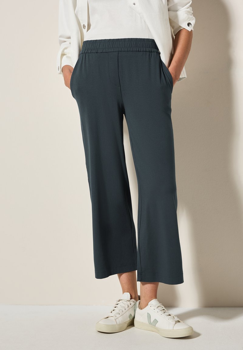 Pantalons cropped vert foncé avec une taille élastique et des poches, fabriqués en tissu lisse. Assortis à des baskets blanches avec des accents verts.