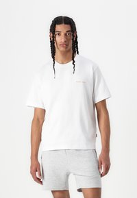 Camiseta de algodón blanca con mangas cortas, que presenta un pequeño logo naranja en el pecho. Combinada con pantalones cortos grises, el modelo está de pie frente a un fondo simple.