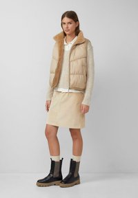 Vrouw in een beige pufferjack met bontvoering, crème trui, beige rok en zwarte chunky laarzen, staande tegen een effen grijze achtergrond.