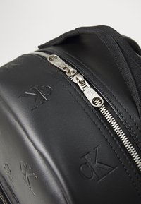 Zaino in ecopelle nera con logo impresso, hardware della cerniera in argento e dettagli in tessuto testurizzato. Presenta una forma strutturata e una maniglia imbottita.