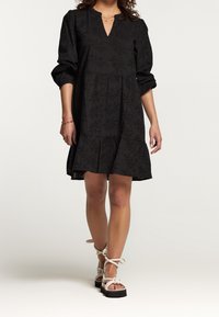 Femme portant une robe noire texturée à manches longues avec un ourlet à volants et des sandales à plateforme blanches avec des brides autour de la cheville, se tenant sur un fond blanc.