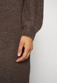 Pull en maille brune mettant en valeur une texture douce ; gros plan sur une main près de la manche, soulignant les bords c ôtelés et la couleur riche.