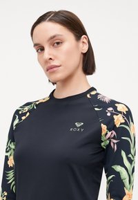 Čierny dlhý rukáv rash guard s kvetinovým vzorom na rukávoch, zelené logo Roxy na prednej strane a hladká, elastická látka.