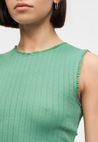 Gros plan d'une personne portant un haut côtelé vert sans manches avec de petites bordures à volants le long de l'encolure et des emmanchures, portant des boucles d'oreilles argentées en forme d'anneau.