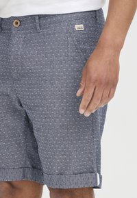 Homme portant un short bleu à motifs avec les ourlets retroussés et une chemise blanche, la main reposant près de la poche du short.