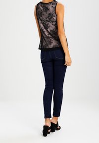 Top senza maniche trasparente nero con motivo in pizzo floreale, abbinato a jeans skinny blu scuro e scarpe con tacco nere.