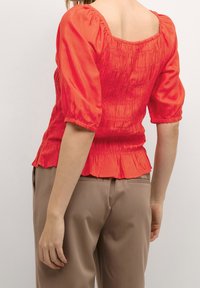 Top rosso a maniche corte con arricciatura elastica e scollatura quadrata; dettagli arricciati e orlo svasato, abbinato a pantaloni beige.