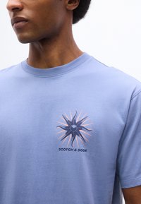 Άνδρας που φοράει ανοιχτό μπλε μπλουζάκι με μοτίβο σκοτεινού μπλε και πορτοκαλί ηλιακής ακτίνας και το λογότυπο "SCOTCH & SODA" στο στήθος, στραμμένος προς τα αριστερά.