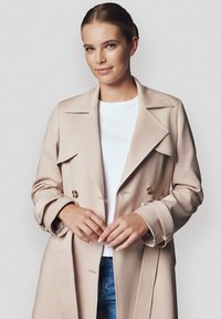 Beżowy podwójnie zapięty płaszcz trench z dużymi klapami i złotymi guzikami, uszyty z gładkiego materiału. Nosi się go na białym t-shircie i dżinsach.