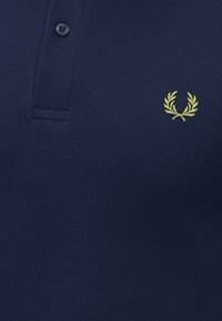 Polo bleu marine avec un bouton noir et un logo de couronne de laurier jaune brodé sur la poitrine.