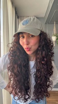 Jeune femme aux longs cheveux bouclés porte une casquette beige et une chemise blanche, faisant une moue joueuse à l'intérieur près d'une fenêtre et d'une plante suspendue.