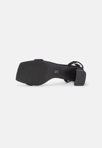 Sort højhælet sandal med en tekstureret sål, justerbar ankelrem og en rund tådesign. Materialet virker blødt og velouragtigt.