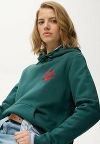 Groene hoodie met een voorvak, capuchon met trekkoord en een rode geborduurde afbeelding. Draagt over een blauw-wit gestreept shirt.