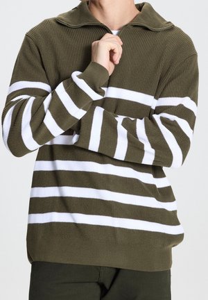 Mann in olivgrüner Strickjacke mit weißen horizontalen Streifen, Reißverschlusskragen und dunkler Hose, posierend mit der Hand nahe am Hals.