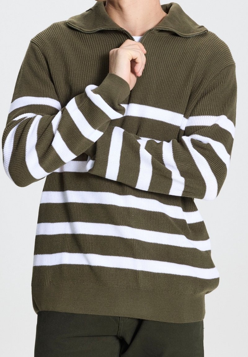 Hombre con suéter de punto verde oliva con rayas horizontales blancas, cuello con cremallera y pantalones oscuros, posando con la mano cerca del cuello.