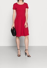 Punainen lyhythihainen mekko, jossa on tiukka yläosa ja laskostettu hame, valmistettu pehmeästä kankaasta. Yhdistetty mustiin korkokenkiin ja teksturoituun mustaan laukkuun.