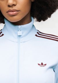 Veste à fermeture éclair bleu clair avec col haut, avec des rayures bordeaux sur les épaules et un petit logo bordeaux sur la poitrine. Texture lisse.