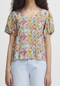 Blouse à motifs colorés avec un col en V, manches courtes bouffantes, présentant des motifs en forme de losange dans des teintes de rose, orange, turquoise et jaune.