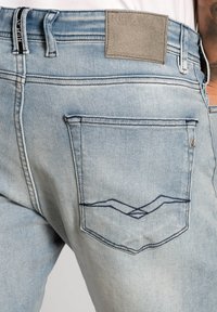 Jeans din denim de culoare albastră deschisă au o pungă pe spate cu un detaliu de cusătură în formă de V și o etichetă rectangulară din piele la talie.