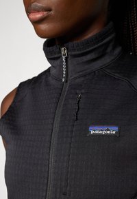 Κοντινό πλάνο ενός ατόμου που φοράει μαύρο αμάνικο ζακέτα Patagonia με φερμουάρ, ψηλό γιακά και φερμουάρ σε τσέπη στο στήθος.