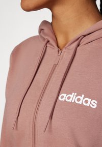 Rózsaszín, cipzáros kapucnis pulóver, puha anyagból, elöl fehér "adidas" logóval. Láthatók a húzózsinórok és a textúrás részletek.