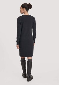 Schwarzes langärmliges Kleid mit schlichter Silhouette. Weicher Strickstoff, knielang, kombiniert mit hohen schwarzen Stiefeln und grauen Socken.