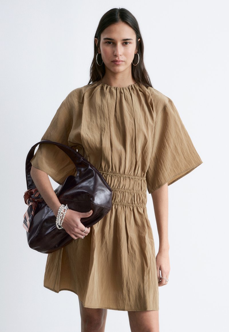 Femme portant une robe ample marron clair jusqu'au genou, tenant un grand sac en cuir marron foncé avec un foulard noué, portant des boucles d'oreilles créoles et des bracelets en perles.