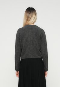 Woman with long blonde hair wearing a dark gray sweater and black pleated skirt, standing facing a light gray wall.
