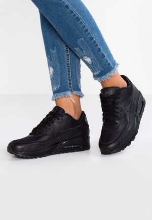 Černé tenisky Nike Air Max s texturovaným svrškem, šněrováním a viditelným vzduchovým polstrováním. Nosič má na sobě roztrhané džínové kalhoty.